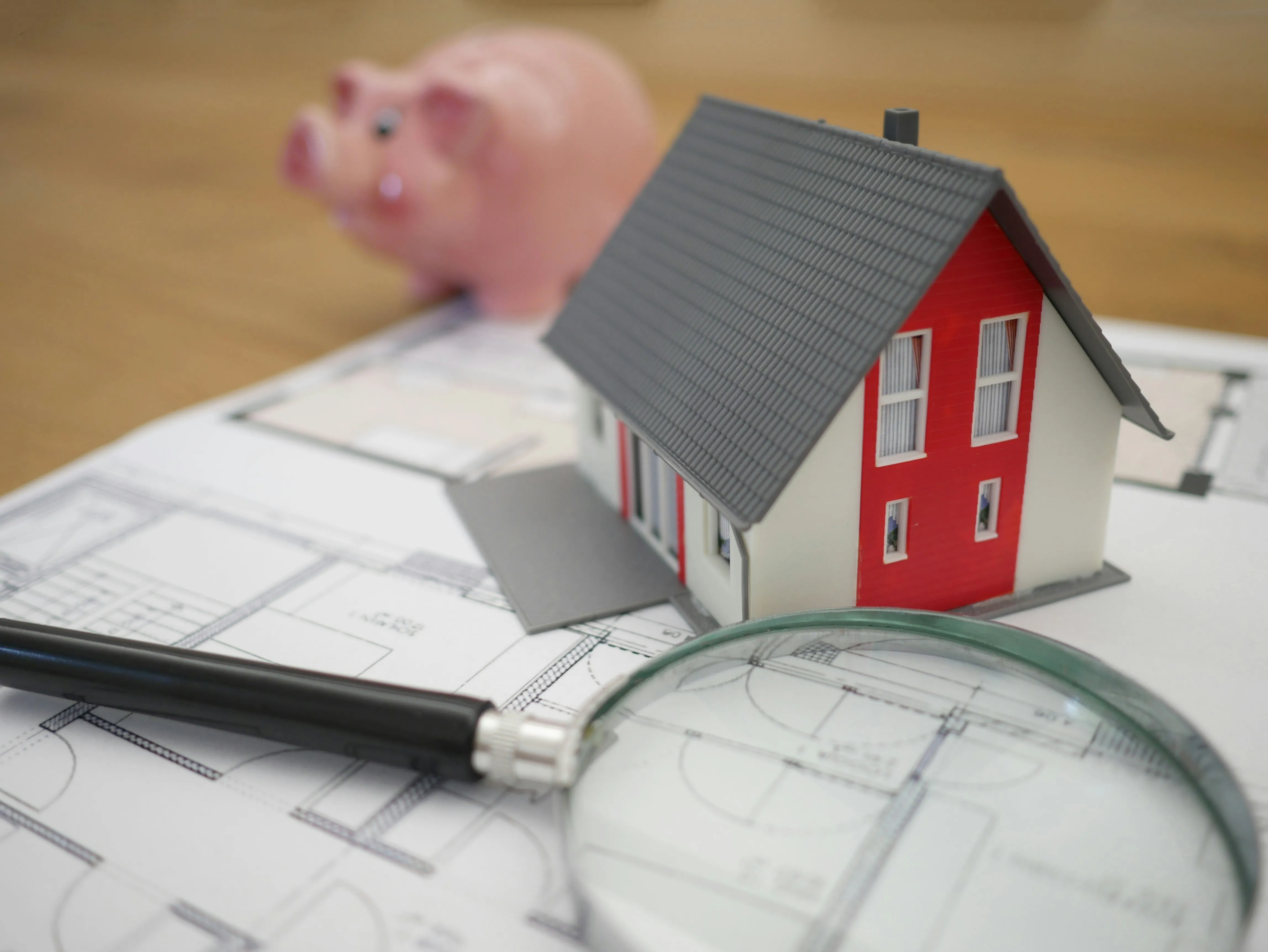 house and magnifying glass on top of blueprints with a piggy bank in the background